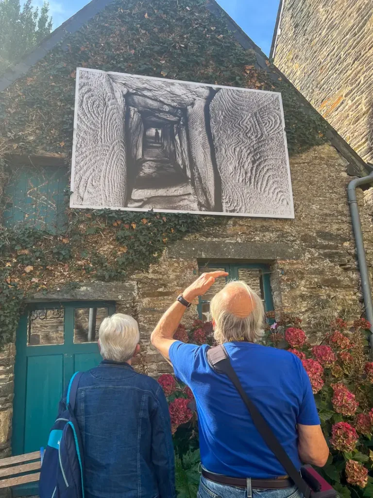 un couple regarde le tirage photo du mégalithe sur le mur d'une maison du village